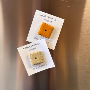 An orange cheez it refrigerator magnet and a yellow white cheddar cheezit magnet mounted on white cardstock are on a stainless steel refrigerator background.