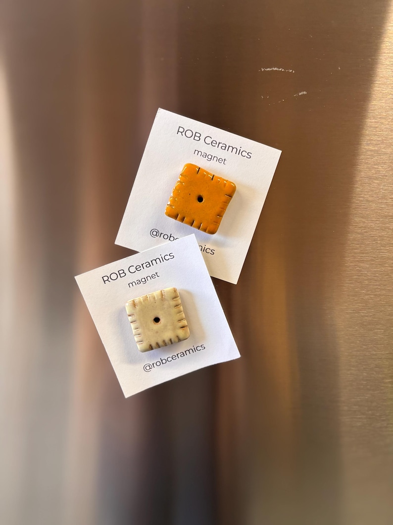 An orange cheez it refrigerator magnet and a yellow white cheddar cheezit magnet mounted on white cardstock are on a stainless steel refrigerator background.