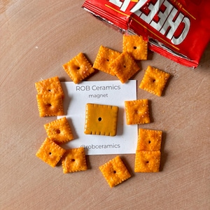 May include: A ceramic magnet shaped like a Cheez-It cracker. The magnet is orange and has a small hole in the center. The magnet is on a white card with the text "ROB Ceramics magnet @robceramics".