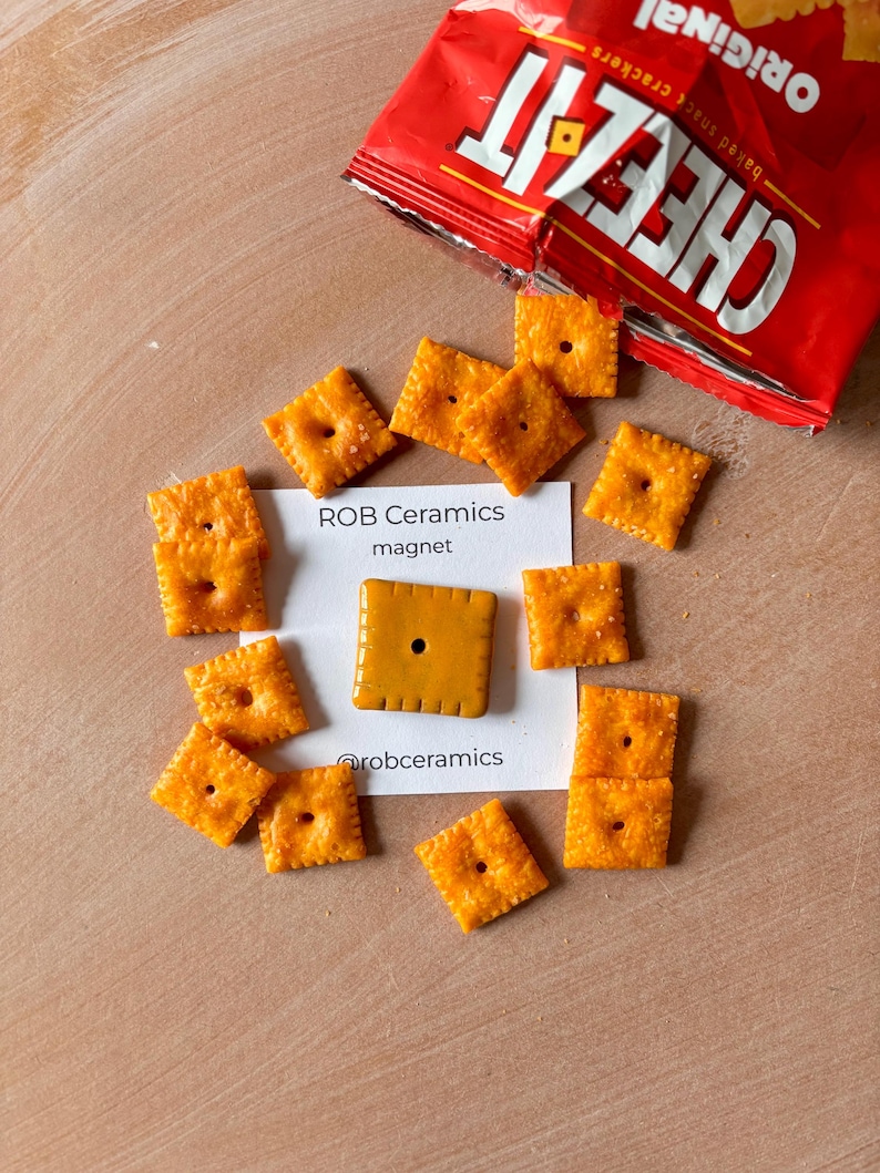 May include: A ceramic magnet shaped like a Cheez-It cracker. The magnet is orange and has a small hole in the center. The magnet is on a white card with the text "ROB Ceramics magnet @robceramics".