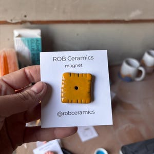 An orange cheez it refrigerator magnet mounted on white cardstock is held by a hand.