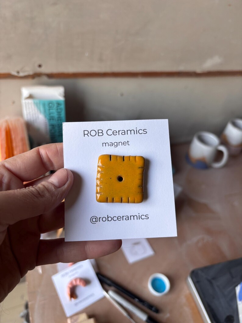 An orange cheez it refrigerator magnet mounted on white cardstock is held by a hand.