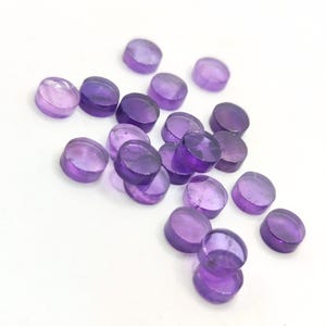 May include: A scattering of small, round, amethyst-coloured gemstones on a white surface. The gemstones have a translucent appearance, with varying shades of purple and a polished finish. The arrangement is random, with some stones overlapping.
