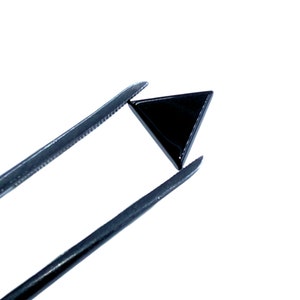 Pode incluir: Uma pequena gema triangular preta é segurada por uma pinça contra um fundo branco. A gema tem uma superfície polida e reflexiva. A pinça é cinza escuro.