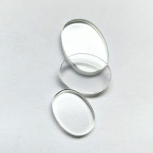 Puede incluir: Tres cristales de reloj de vidrio transparente de diferentes tamaños y formas, dos ovalados y uno redondo, sobre un fondo blanco.