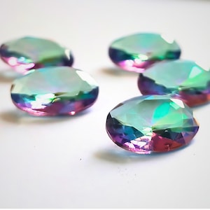 May include: Five iridescent, oval gemstones with cut facets. The gems reflect light, displaying a spectrum of colours including green, blue, purple, and pink. They are scattered on a white surface, creating a sparkling effect.
