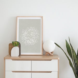 K&ouml;nnte beinhalten: Ein gerahmter Druck einer wei&szlig;en Linienzeichnung einer Blume auf hellgrauem Hintergrund. Der Druck befindet sich auf einer wei&szlig;en Kommode mit einer kleinen Sukkulente in einem wei&szlig;en Topf und einer wei&szlig;en Mondlampe auf einem Holzst&auml;nder.