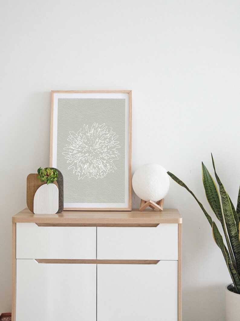 K&ouml;nnte beinhalten: Ein gerahmter Druck einer wei&szlig;en Linienzeichnung einer Blume auf hellgrauem Hintergrund. Der Druck befindet sich auf einer wei&szlig;en Kommode mit einer kleinen Sukkulente in einem wei&szlig;en Topf und einer wei&szlig;en Mondlampe auf einem Holzst&auml;nder.