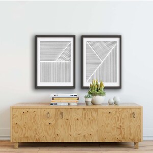 May include: Two black framed prints with abstract line art designs on a white background. The prints are hanging on a white wall above a light wood cabinet with three doors.