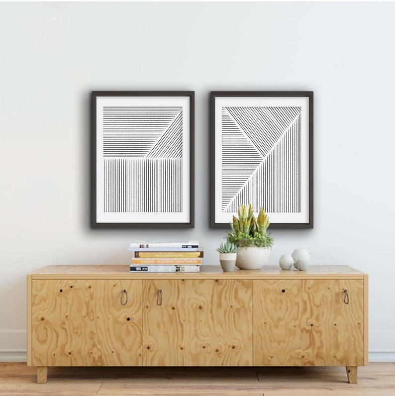 May include: Two black framed prints with abstract line art designs on a white background. The prints are hanging on a white wall above a light wood cabinet with three doors.