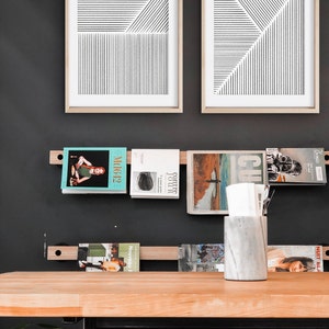 May include: A wooden shelf with black metal brackets holds a collection of magazines and a white marble vase. Two framed black and white line art prints hang on the wall behind the shelf. The shelf is mounted on a black wall.