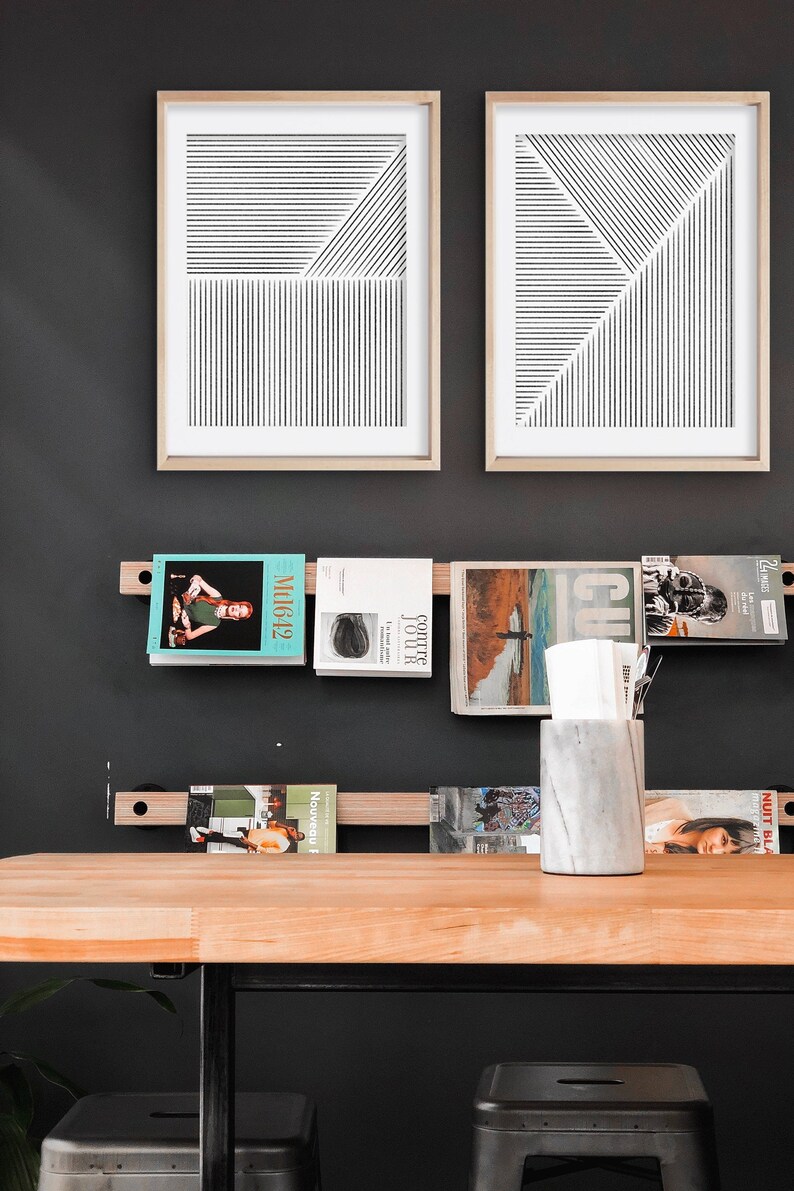 May include: A wooden shelf with black metal brackets holds a collection of magazines and a white marble vase. Two framed black and white line art prints hang on the wall behind the shelf. The shelf is mounted on a black wall.
