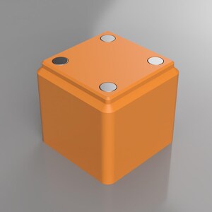 May include: An orange cube-shaped object with four silver circular accents on the top surface. The object has a smooth, matte finish and rounded corners. The background is a solid, neutral gray.