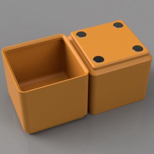 May include: An open, orange square box with rounded corners sits next to a closed, orange square box. The closed box has four black circular magnets on its top surface. The boxes are on a gray surface.