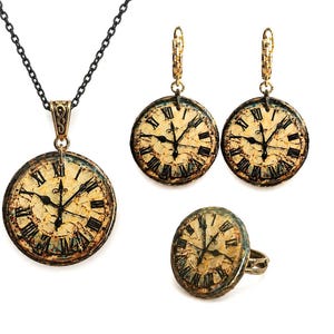 May include: A jewellery set comprising a necklace, earrings, and a ring, each with a vintage clock face design. The necklace has a black chain and a gold-toned pendant. The earrings and ring also have gold-toned accents. The clock faces feature black Roman numerals.