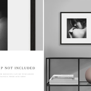 May include: A black and white photograph of a woman is framed in a black frame and hung on a grey wall. The frame is on a black shelf with a white book and a round, light pink vase.