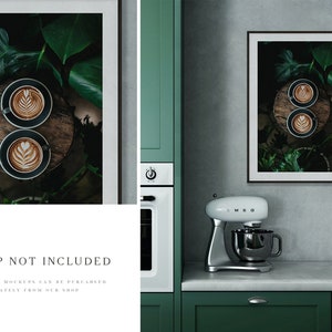 May include: Two cups of coffee with latte art on a wooden surface surrounded by green leaves in a black frame hanging on a wall in a kitchen setting.