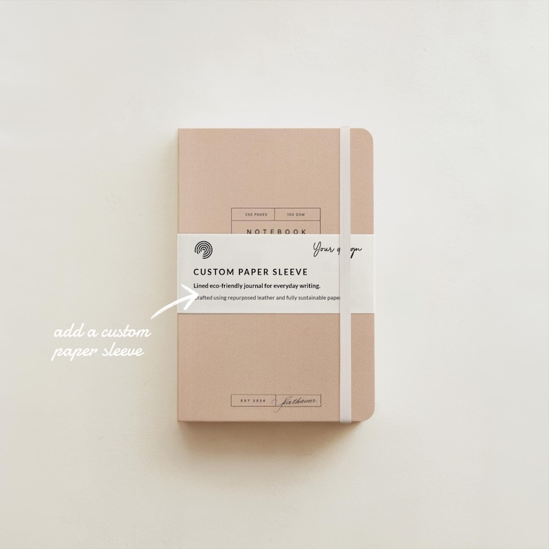 May include: A light brown notebook with a white elastic band closure and a white label that says "CUSTOM PAPER SLEEVE" with the text "Lined eco-friendly journal for everyday writing. Crafted using repurposed leather and fully sustainable paper."