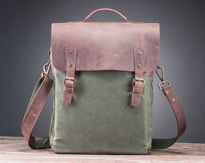 May include: A green canvas and brown leather messenger bag with a top handle and adjustable shoulder strap. The bag has two buckles on the front and a metal clasp on the shoulder strap.