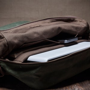 May include: A green canvas messenger bag with a brown leather strap and a zipper closure. The bag is open and a white laptop and a cell phone are inside.