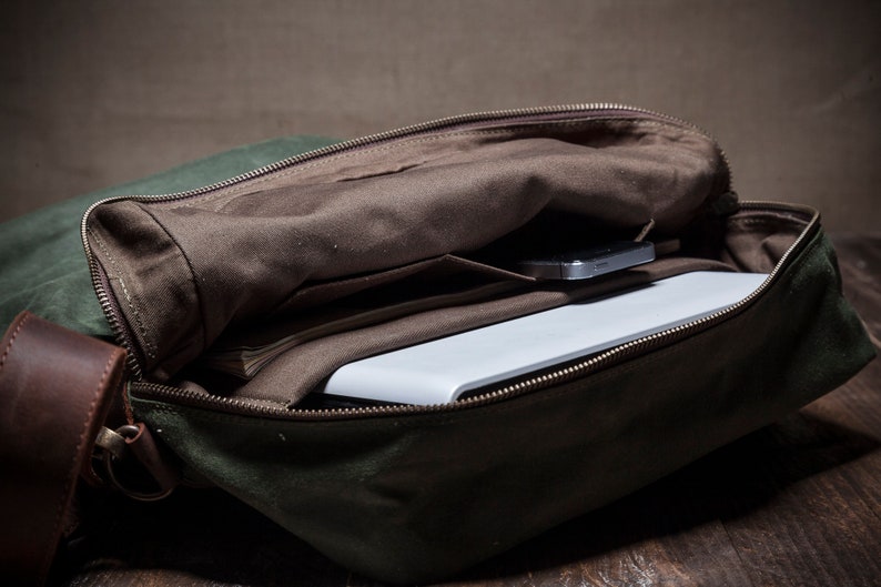 May include: A green canvas messenger bag with a brown leather strap and a zipper closure. The bag is open and a white laptop and a cell phone are inside.