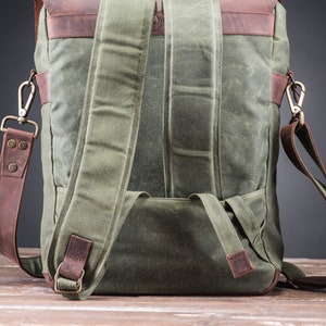 May include: A green canvas backpack with brown leather straps and accents. The backpack has a large main compartment and a smaller front pocket. The straps are adjustable and can be worn on the shoulders or as a crossbody bag.