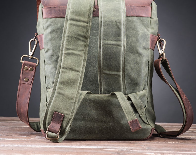 May include: A green canvas backpack with brown leather straps and accents. The backpack has a large main compartment and a smaller front pocket. The straps are adjustable and can be worn on the shoulders or as a crossbody bag.