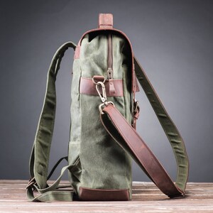 May include: A green canvas backpack with brown leather accents and a zipper closure. The backpack has a top handle and adjustable shoulder straps.