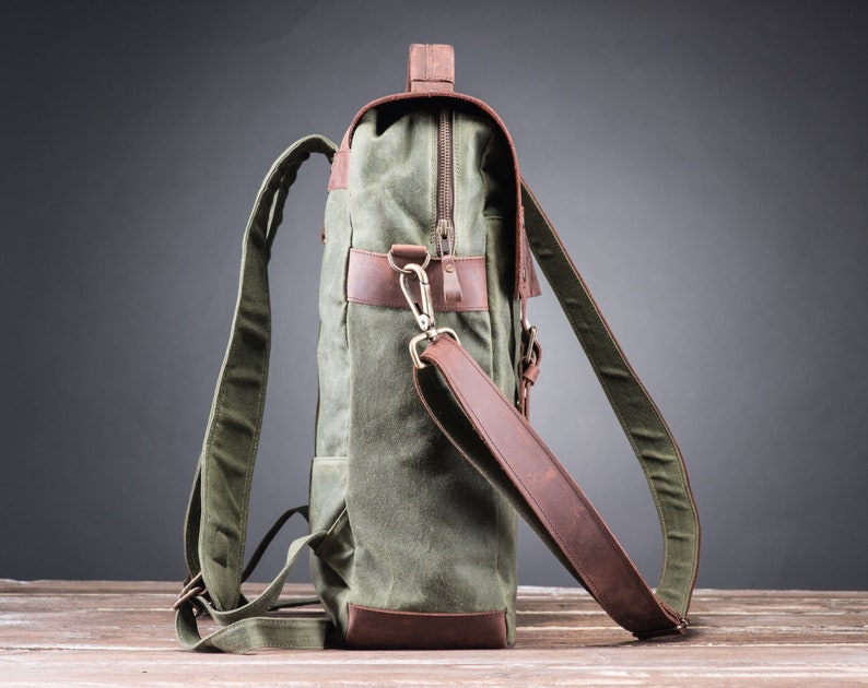 May include: A green canvas backpack with brown leather accents and a zipper closure. The backpack has a top handle and adjustable shoulder straps.