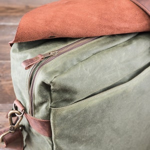 May include: A green canvas bag with a brown leather zipper pull and a brown leather flap. The bag has a brown leather strap and a brass clasp.