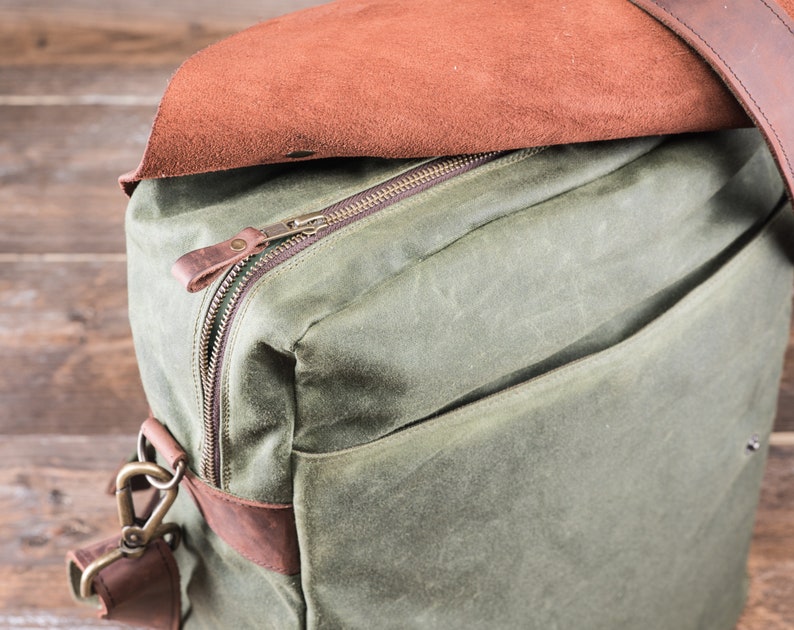 May include: A green canvas bag with a brown leather zipper pull and a brown leather flap. The bag has a brown leather strap and a brass clasp.