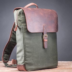 Puede incluir: Una mochila de lona verde con una solapa de cuero marrón y cierre de hebilla. La mochila tiene correas ajustables y un bolsillo con cremallera en la parte delantera.