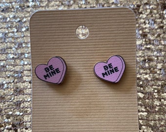 May include: A pair of pink heart-shaped earrings with the text "BE MINE" on each. The earrings are displayed on a brown card.
