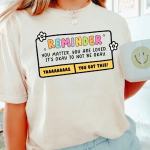 May include: A white t-shirt with a yellow and black reminder box that says "REMINDER YOU MATTER. YOU ARE LOVED. IT'S OKAY TO NOT BE OKAY. YAAAAAAAAS. YOU GOT THIS!"