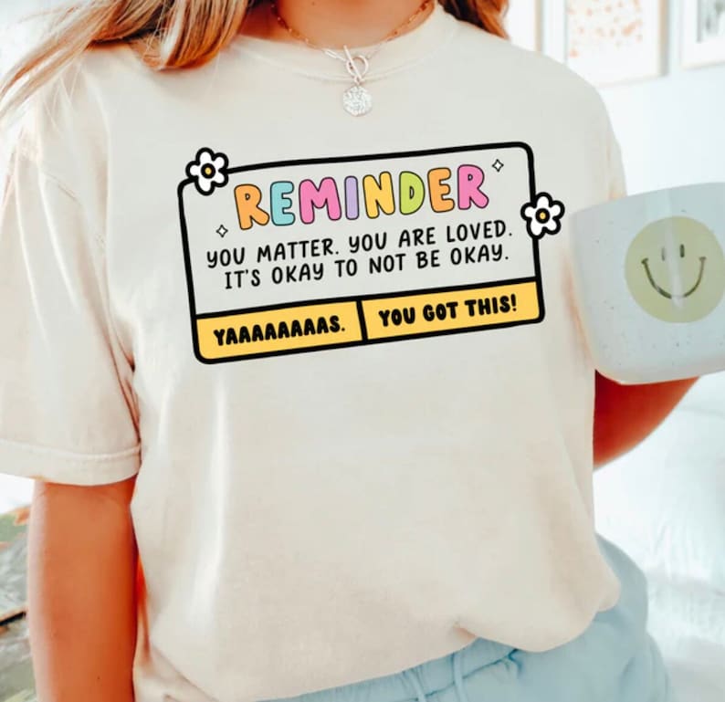 May include: A white t-shirt with a yellow and black reminder box that says "REMINDER YOU MATTER. YOU ARE LOVED. IT'S OKAY TO NOT BE OKAY. YAAAAAAAAS. YOU GOT THIS!"