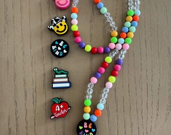 Puede incluir: Un cordón de cuentas de colores con una variedad de dijes. El cordón presenta una mezcla de cuentas de colores del arcoíris y cuentas de cristal transparente. Los dijes incluyen una vaquera sonriente, una pila de libros con un gusano y una manzana con el texto "A+ Teacher". El cordón tiene el texto "BE KIND".
