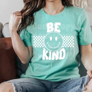 May include: A teal t-shirt with a white checkered pattern and the words "BE KIND" in a retro font with a smiley face in the middle.