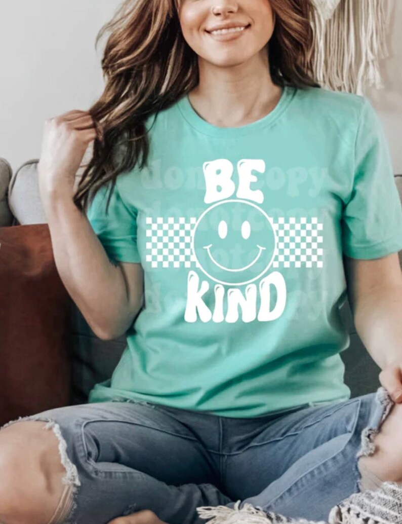 May include: A teal t-shirt with a white checkered pattern and the words "BE KIND" in a retro font with a smiley face in the middle.