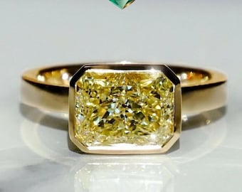 May include: A gold ring featuring a large, radiant-cut yellow diamond. The diamond is set in a bezel setting, and the band is a simple, polished gold. The ring is displayed on a reflective surface.