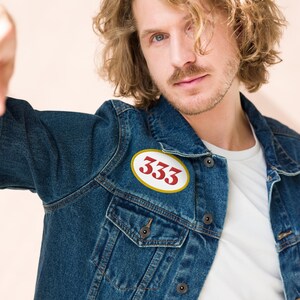 Könnte beinhalten: Ein Mann trägt eine blaue Jeansjacke mit einem weißen ovalen Patch, auf dem "333" in roten Buchstaben steht.