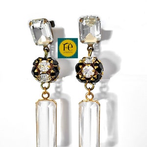 Könnte beinhalten: Ein Paar elegante Ohrringe mit einem klassischen Design. Jeder Ohrring hat einen klaren, rechteckigen Kristallanhänger, eine schwarz-goldene Perlenkugel und einen weiteren rechteckigen Kristall. Die Ohrringe sind in einer goldfarbenen Fassung gehalten. Der Hintergrund ist weiß.