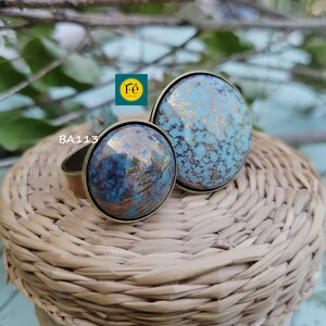 Op de afbeelding: Twee ronde, blauwe en gouden keramische ringen met een gouden metalen band. De ringen liggen op een gevlochten mand.