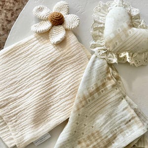 May include: A set of two cream-colored muslin cloths, one plain and one with a tan checkered pattern. A white crocheted flower with a brown center is attached to the plain cloth. A heart-shaped fabric pillow with lace trim sits nearby.