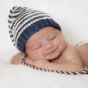Peut inclure: Un nouveau-né portant un bonnet en tricot à rayures bleues et blanches. Le bébé est allongé sur une surface blanche et moelleuse, avec un détail de corde bleu et blanc. Le bébé sourit, les yeux fermés.