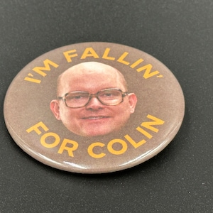 May include: A brown button with a picture of a man with glasses and the text "I'm fallin' for Colin" in yellow.