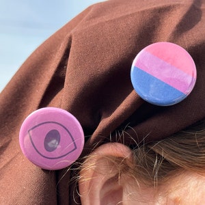 Könnte beinhalten: Zwei runde Pins auf einem braunen Stoffhintergrund. Ein Pin ist rosa, lila und blau mit einem horizontalen Streifenmuster. Der andere Pin ist lila mit einer schwarzen Umrandung eines Auges.