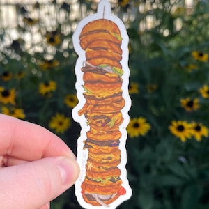 May include: A sticker featuring a tall stack of burgers with various toppings. The burgers are depicted in shades of brown and orange, with visible layers of meat, cheese, lettuce, and onions. The sticker has a white border and is held against a blurred floral background.