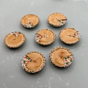 May include: Seven miniature pot pies in silver foil tins, each featuring a golden-brown crust with a wedge cut out to reveal a colorful filling of small green peas, orange carrot cubes, and white potato chunks arranged in a circular pattern.