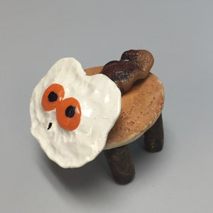 Könnte beinhalten: Ein Keramikhocker mit drei Beinen, bemalt wie ein Pfannkuchen mit einem Spiegelei obenauf und einem Stück Speck an der Seite.