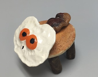 Może przedstawiać: Ceramiczny stołek z trzema nogami, pomalowany tak, aby wyglądał jak naleśnik z jajkiem sadzonym na wierzchu i kawałkiem bekonu z boku.